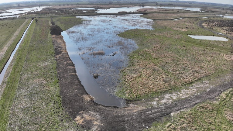 An aerial view of an expansive green, flat landscape, showing bare earth (brown mud) where excavations have recently taken place, and areas of open water shining silver-blue in the spring sun - a muddy bank surrounds the area of open water and a long, straight ditch is located to its side